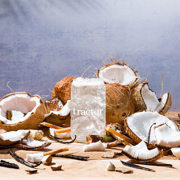 A group of coconuts and a glass of ice on a table with a Tractor Beverage Co. Organic Coconut Beverage bag in box.