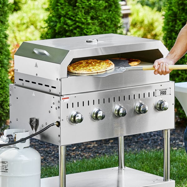 A person using a Backyard Pro pizza oven attachment to cook a pizza on a grill.