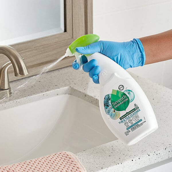 A person in blue gloves spraying Seventh Generation Tub and Tile Cleaner from a white bottle.