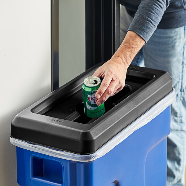 A hand putting a can into a Toter slimline black mixed recycling lid on a trash can.