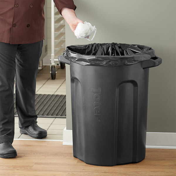 A man in a chef's uniform throwing a white object into a black Toter trash can.