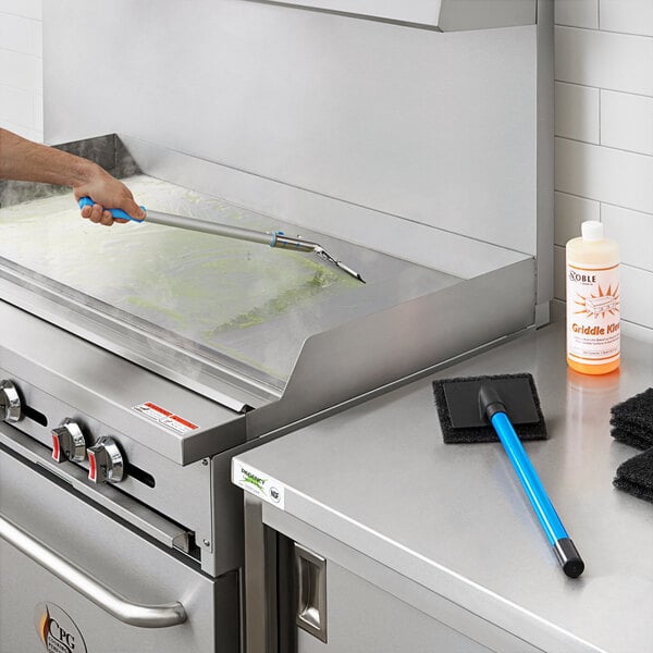 A person using a white and orange Griddle Cleaning Starter Kit tool with a long blue handle to clean a stove top.