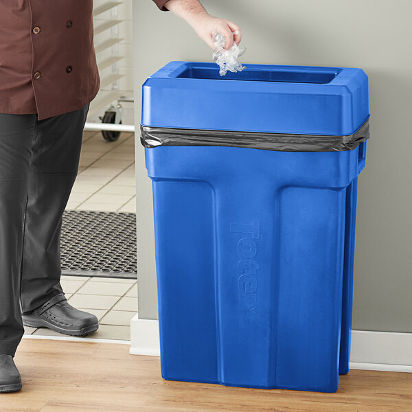 A man standing next to a Toter blue trash can with a blue lid.
