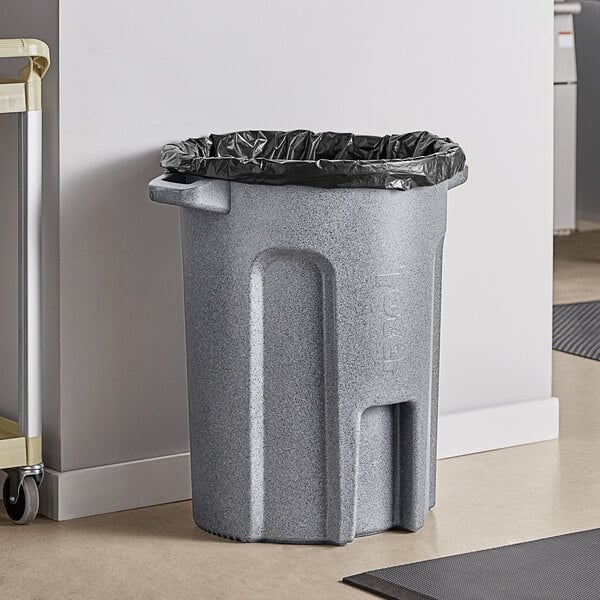 A Toter dark gray granite round trash can with a black bag over it sits in a room in a corporate office cafeteria.