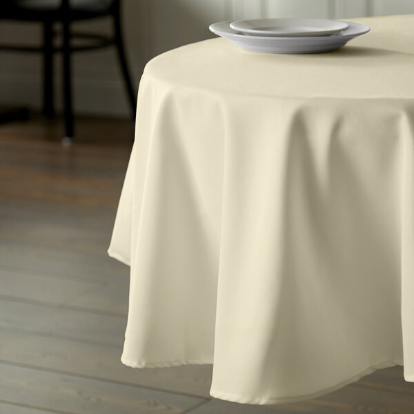 A table with an ivory Intedge round tablecloth on it with a white plate.