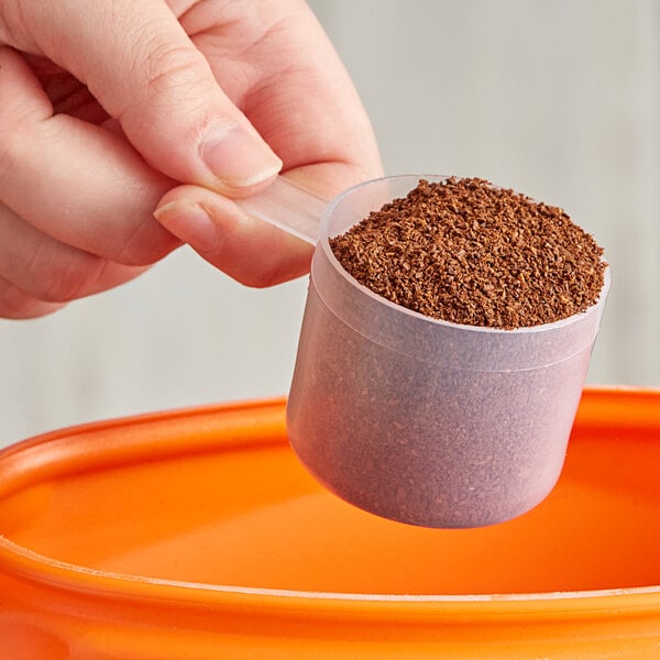 A hand holding a 60 cc Polypropylene scoop of brown powder over a plastic container.