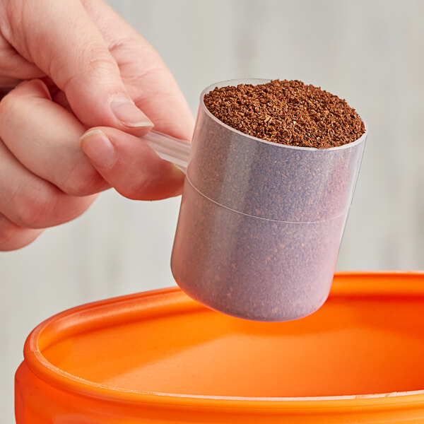 A hand using a polypropylene scoop to fill a measuring cup with brown powder.