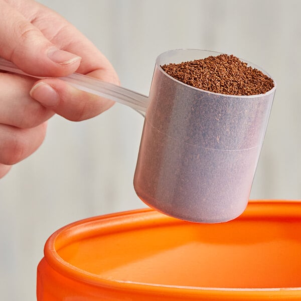 A hand using a long-handled polypropylene scoop to pour brown granules from a measuring cup.