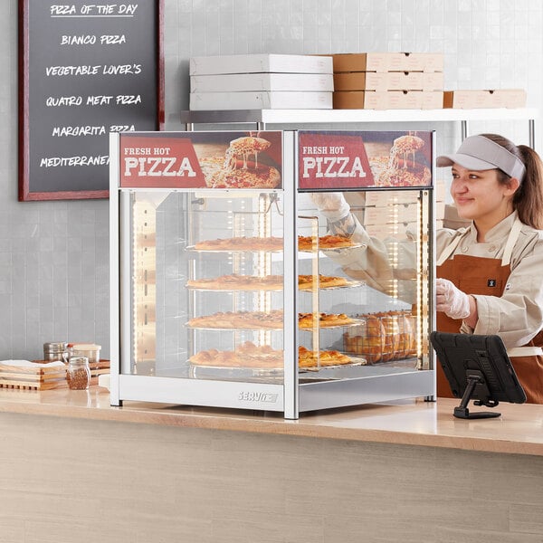 A woman in a brown apron and gloves using a ServIt countertop pizza warmer to display pizzas.