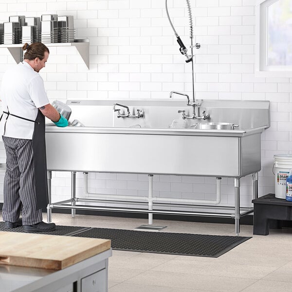 A man in a black apron washing a Regency stainless steel three compartment sink.