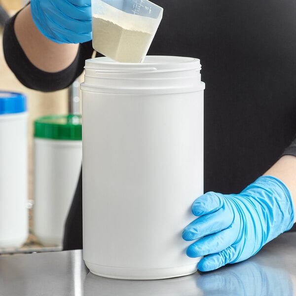 A person in blue gloves pouring white powder into a white HDPE plastic canister.