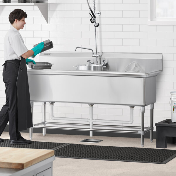 A woman in a black apron using a Regency stainless steel three compartment sink in a professional kitchen.
