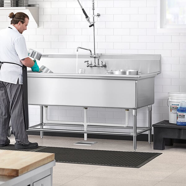 A man in a white shirt and black apron washing dishes in a Regency stainless steel 3 compartment sink.