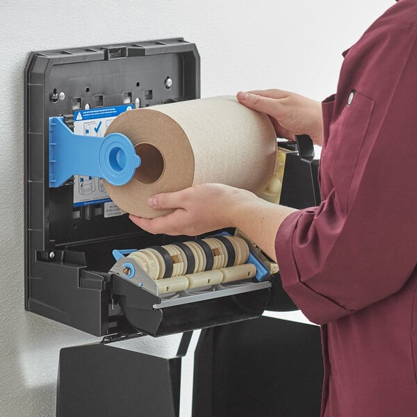 A person putting a Pacific Blue Basic brown paper towel roll into a dispenser.