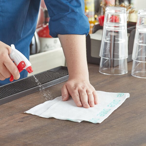 A person using a white and green Dixie foodservice towel to spray a liquid onto a table.