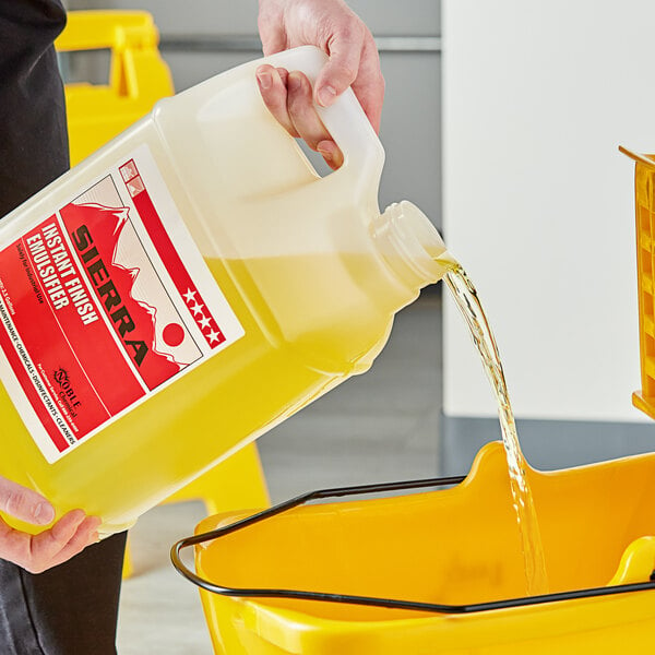 A person pouring Sierra by Noble Chemical concentrated floor finish emulsifier from a yellow jug into a bucket.