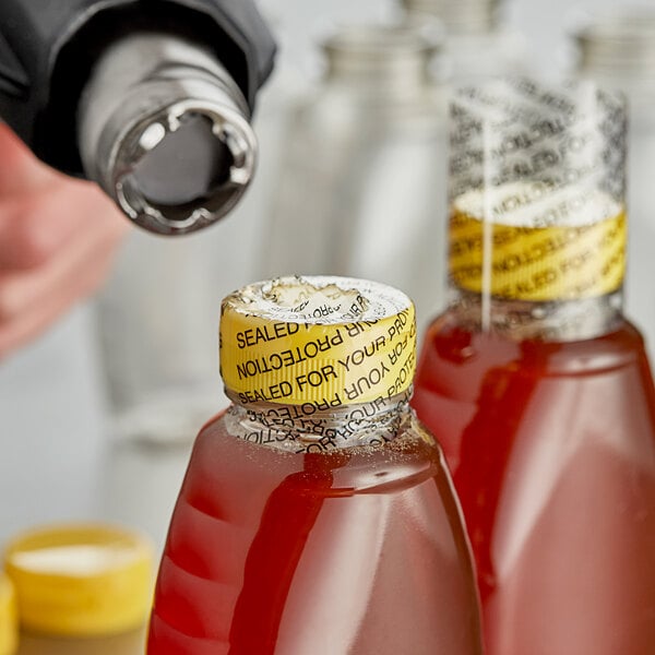 A person using a 2" non-perforated shrink band to seal a bottle of liquid with a yellow cap.