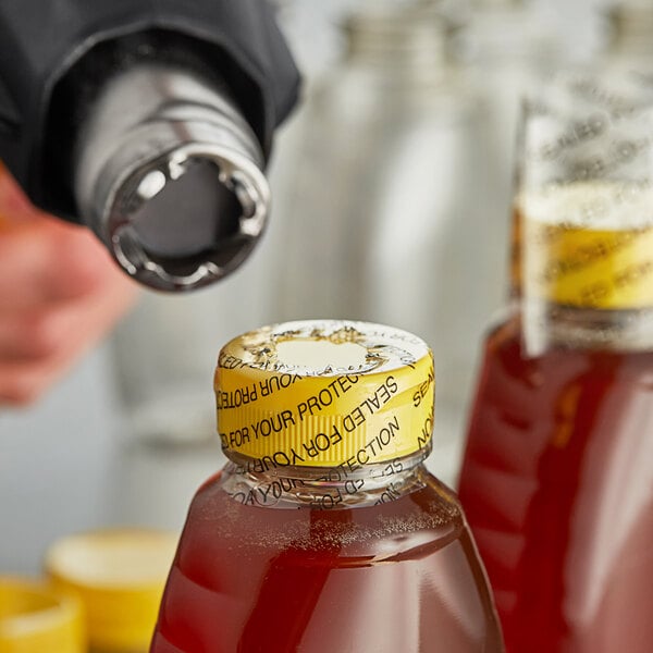 A person using a tool to shrink a 2" perforated shrink band onto a bottle of sauce.