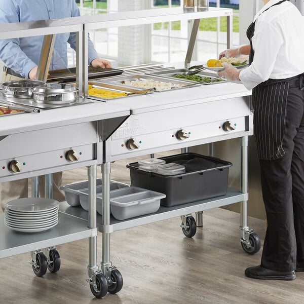 A man and woman using a ServIt three pan open well electric steam table with sneeze guard in a school kitchen.