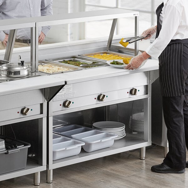 A man in a white shirt and black pants standing next to a ServIt three pan electric steam table on a counter in a school kitchen.