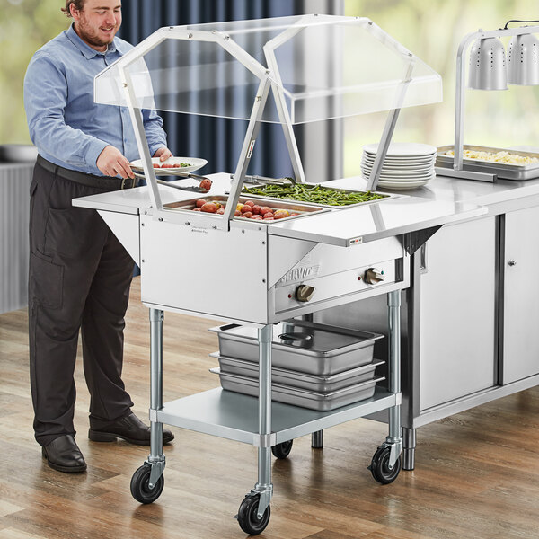 A man standing by a ServIt electric steam table with sneeze guard filled with food.