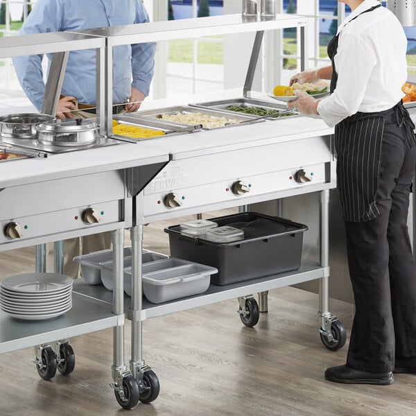 A woman and a man standing in front of a ServIt steam table with clear plastic lids on containers.