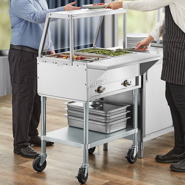 A man and woman standing next to a ServIt open well steam table with sneeze guard.