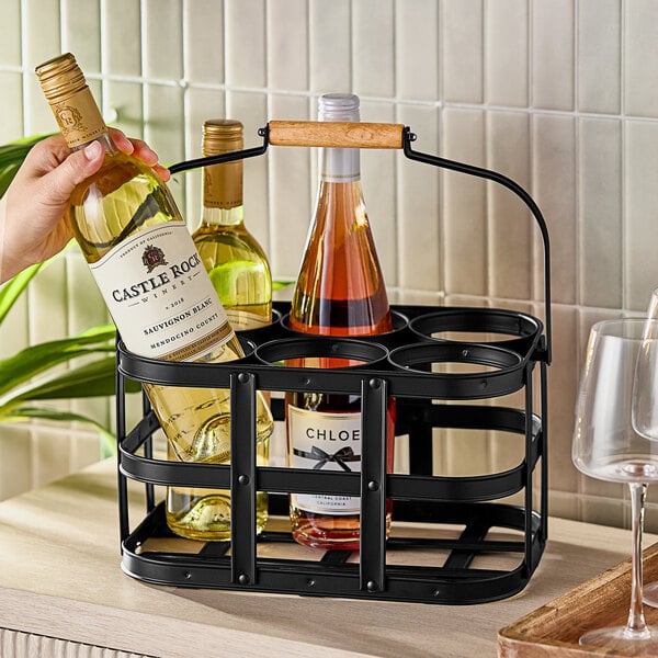 A hand holding a bottle of wine in a Franmara black metal wine bottle caddy on a table.