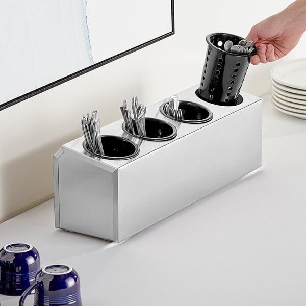 A hand holding a stainless steel flatware organizer with black plastic cylinders filled with utensils.