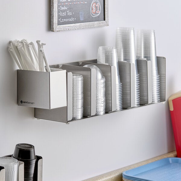 A stainless steel wall-mounted or countertop cup and lid holder with five adjustable sections and an attached straw caddy.