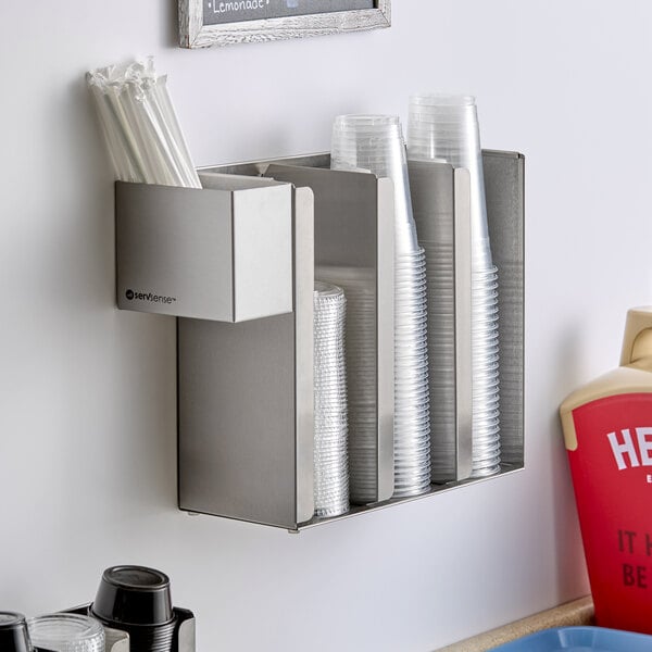 A stainless steel wall-mounted or countertop holder with three sections for cups or lids and an attached straw caddy.