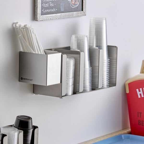 A stainless steel wall-mounted or countertop holder with three sections for cups or lids and an attached straw caddy.