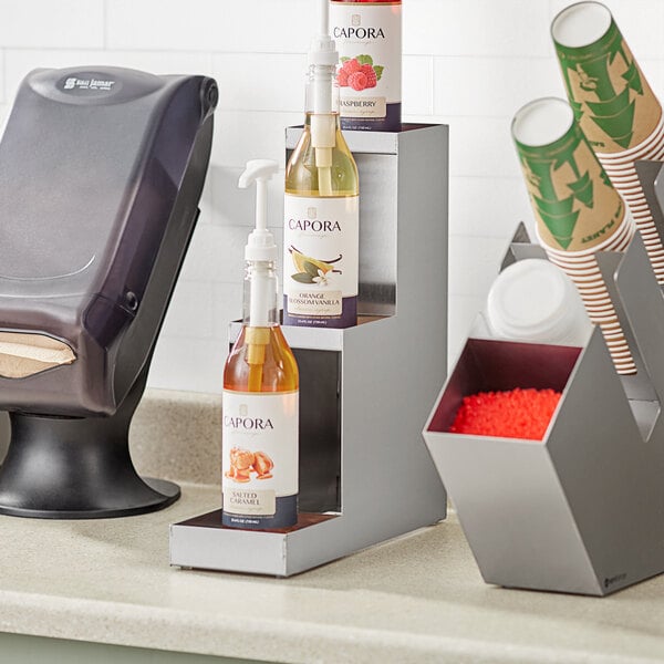 An Acopa stainless steel bottle riser holding three bottles on a counter.