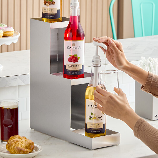 A three-tier stainless steel bottle riser holding syrup bottles for beverage flavoring.