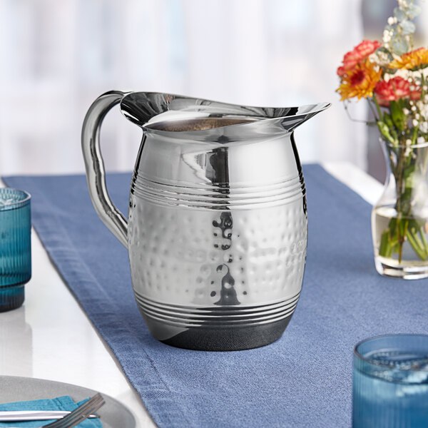 An American Metalcraft hammered stainless steel water pitcher with a silver handle on a table.