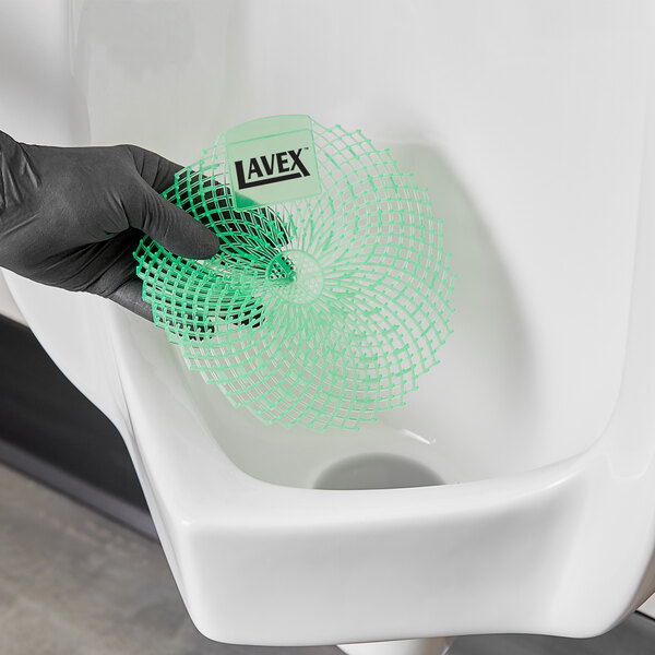 A hand in a black glove holding a Lavex Melon Scented green urinal screen over a urinal.