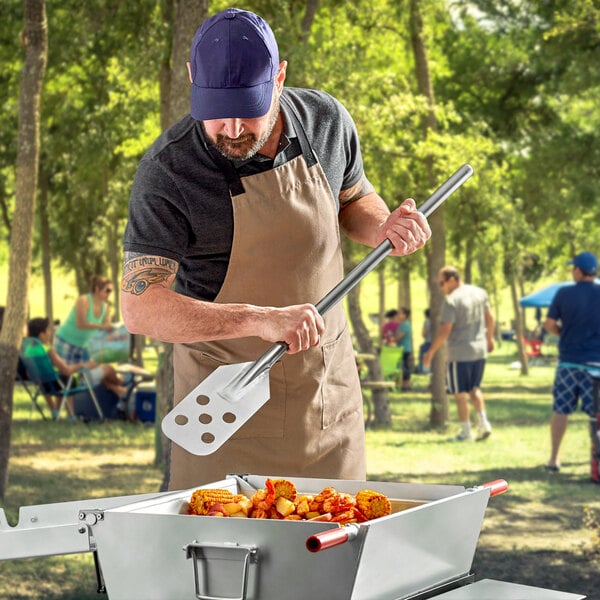 A man in an apron using a Fourt&#233; Perforated Stainless Steel Paddle to cook corn on the cob on a grill.