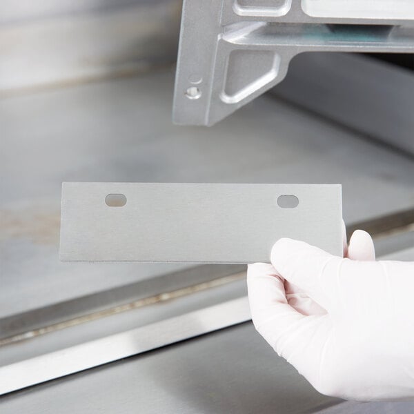 A gloved hand holding a 6" metal grill scraper blade.