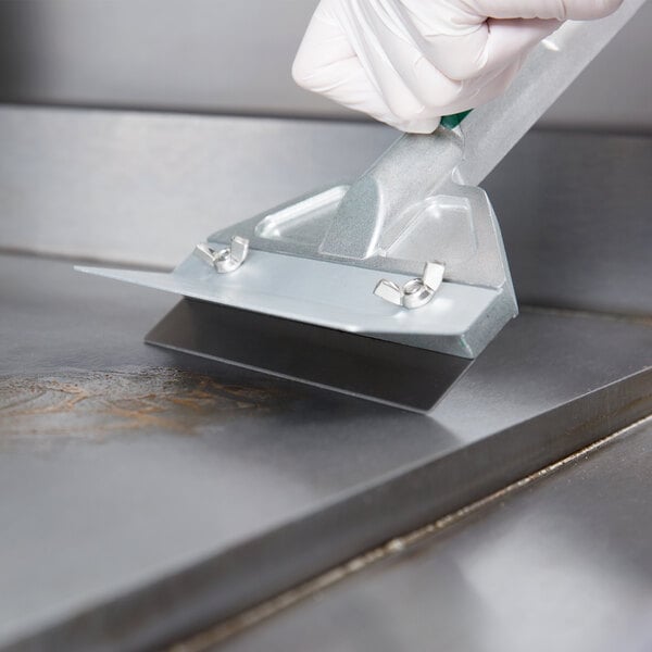 A gloved hand using a 6" metal scraper to clean a metal surface.