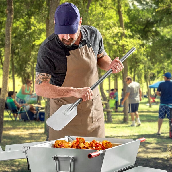 A man in an apron using a Fourt&#233; stainless steel paddle to cook corn on the cob on a grill.