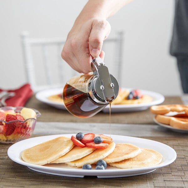 A person using a Tablecraft modern glass syrup dispenser to pour syrup onto a stack of pancakes.