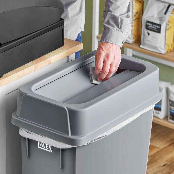 A person using a Lavex gray slim rectangular trash can drop shot lid to throw away paper on a counter.
