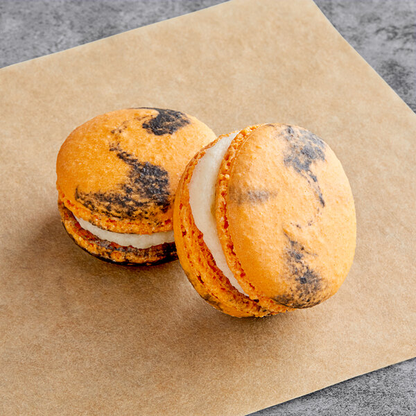 Two Macaron Centrale Pumpkin Cheesecakes with orange and black icing on a piece of paper.