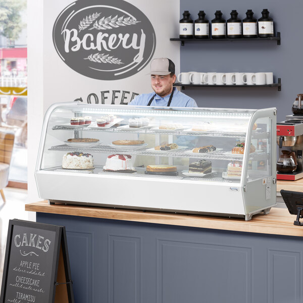 An Avantco white refrigerated countertop bakery display case filled with cakes and cupcakes with a man standing behind it.