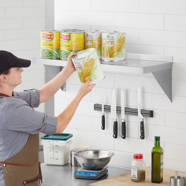 A woman in a brown apron and hat holding a can of green beans on a Regency stainless steel wall shelf.