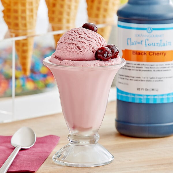 A glass of pink ice cream with cherries and a spoon next to a bottle of LorAnn Black Cherry Flavor Fountain syrup.