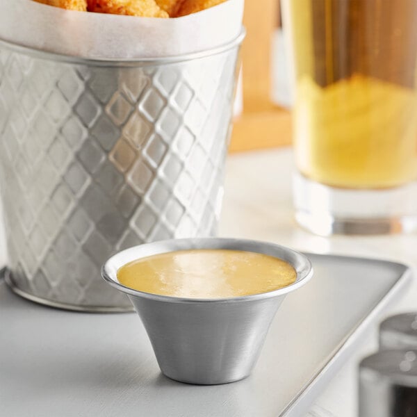 A stainless steel sauce cup filled with sauce on a table next to a tray of chicken nuggets.