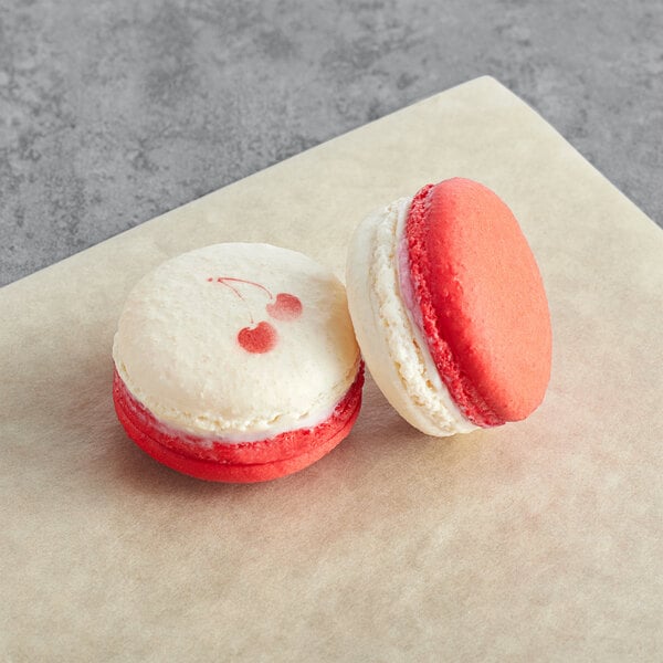 A pink and white Macaron Centrale with cherry icing and a white Macaron Centrale with cherry icing on a table.