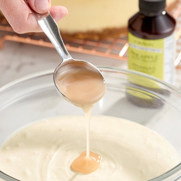 A spoonful of pineapple bakery emulsion being poured into a bowl of batter, with a bottle of LorAnn Oils Pineapple Bakery Emulsion in the background.