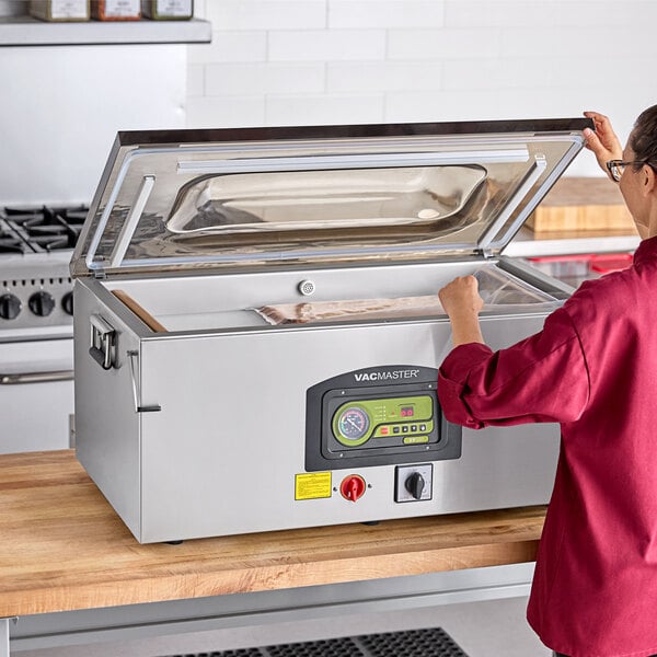 A stainless steel ARY VacMaster VP330 chamber vacuum packaging machine with three seal bars, shown on a kitchen counter with its lid open.